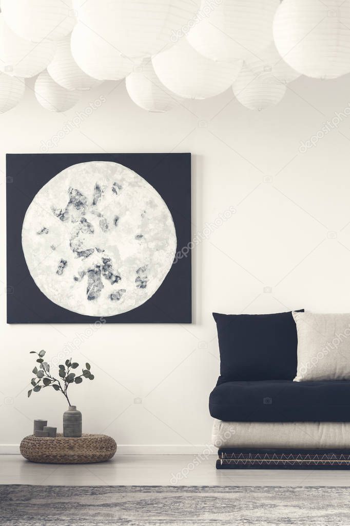 Moon poster above plant on pouf in white flat interior with black sofa and grey carpet. Real photo
