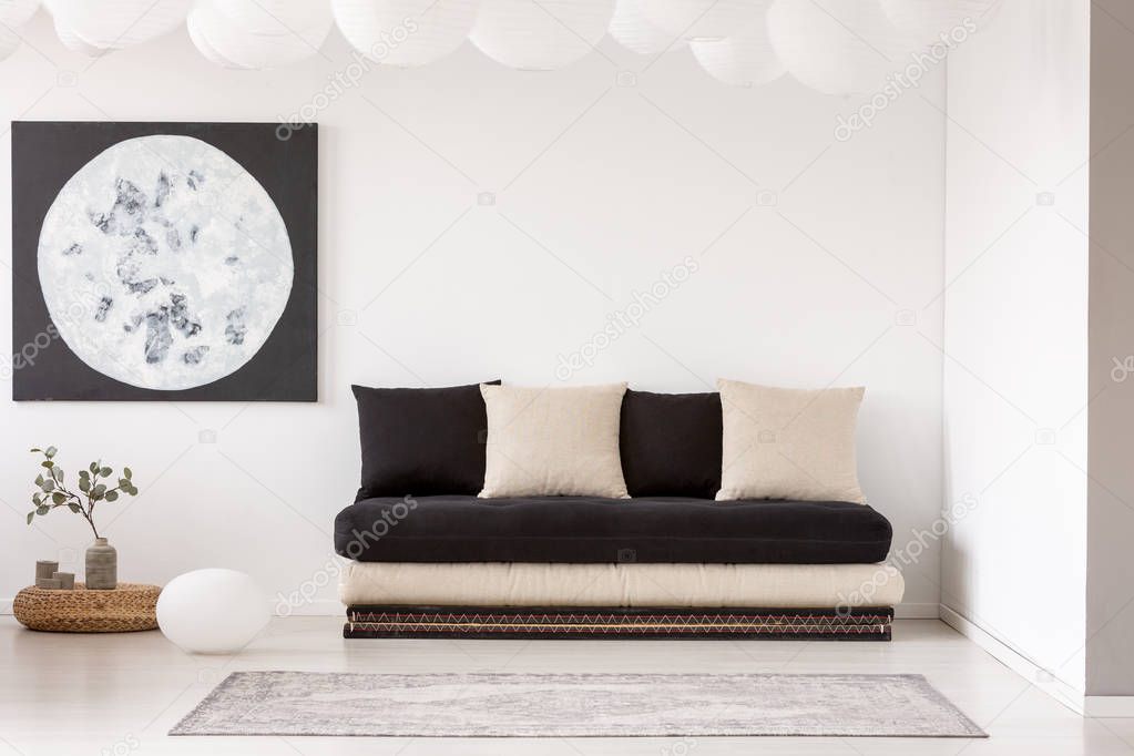 Pillows on black sofa near carpet in white living room interior with plant and moon poster. Real photo