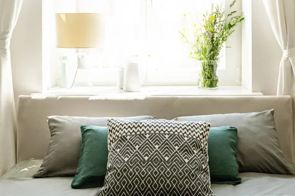 在床的后面用灰色和绿色的枕头对着一个阳光明媚的窗户 用一盏灯和一束野花在一个自然卧室内部的窗台上 真实照片 — 图库照片