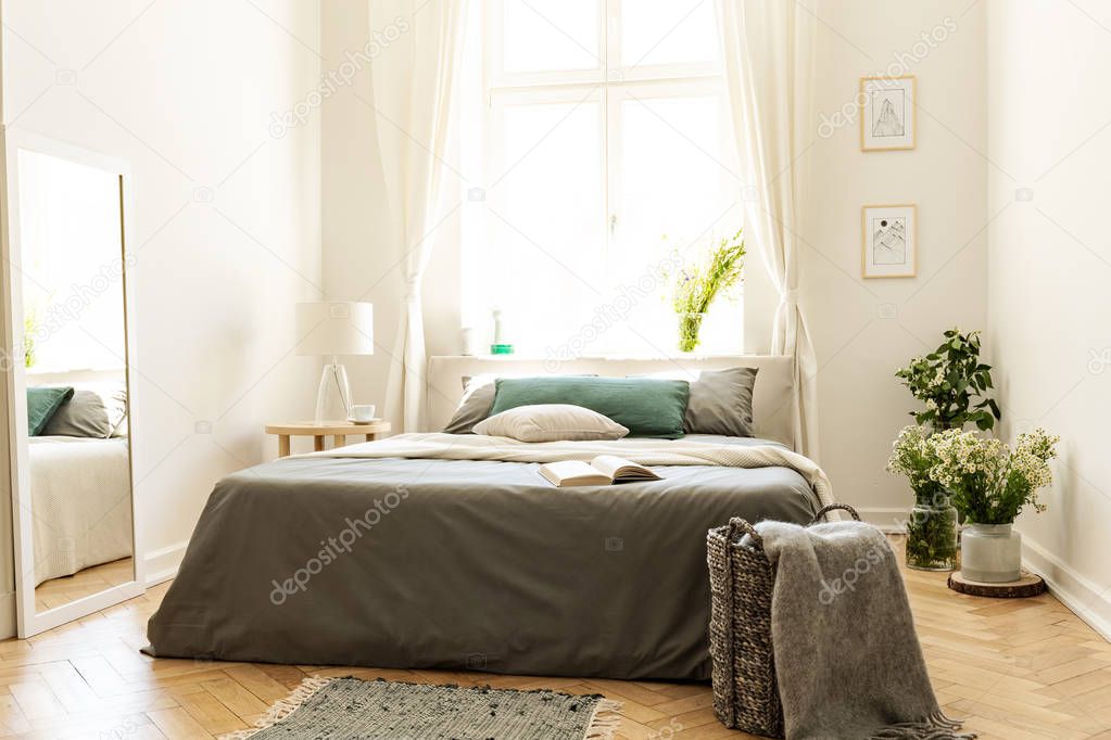 Bright bedroom interior with green decor of a cushion and bunches of wild flowers. Big bed against a sunny window in a villa. Real photo.