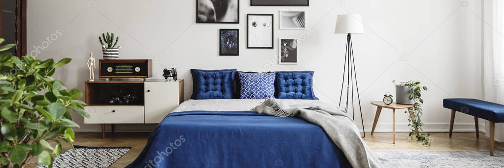 Real photo of an eclectic bedroom interior with graphics on a wall, blue pillows and radio on a cupboard