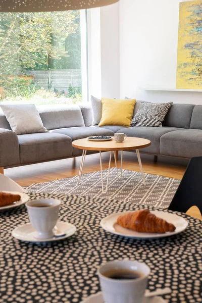 Focus on a gray corner sofa by a big window and a round table and blurry foreground with pastries and coffee in a modern coffee shop interior