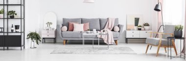 Real photo of white living room interior with grey sofa with cushions, fresh plants and cupboards with decor