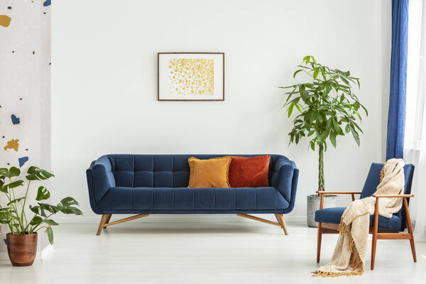 Современное кресло середины века с одеялом и большим диваном с красочными подушками в просторной гостиной с зелеными растениями и белыми стенами. Настоящее фото
.