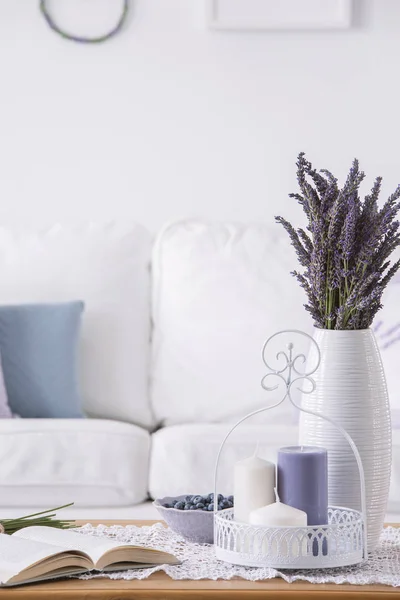 Violet flowers on table with book and candles in white living room interior with sofa. Real photo