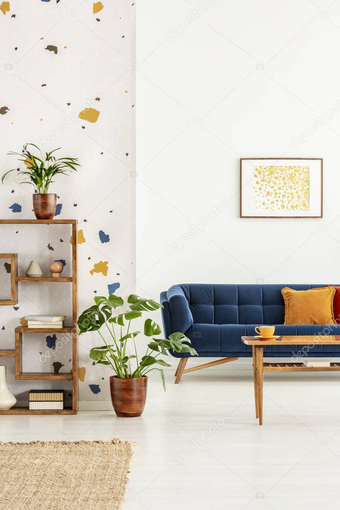 Plants in white apartment interior with poster above blue settee with orange pillow and table. Real photo