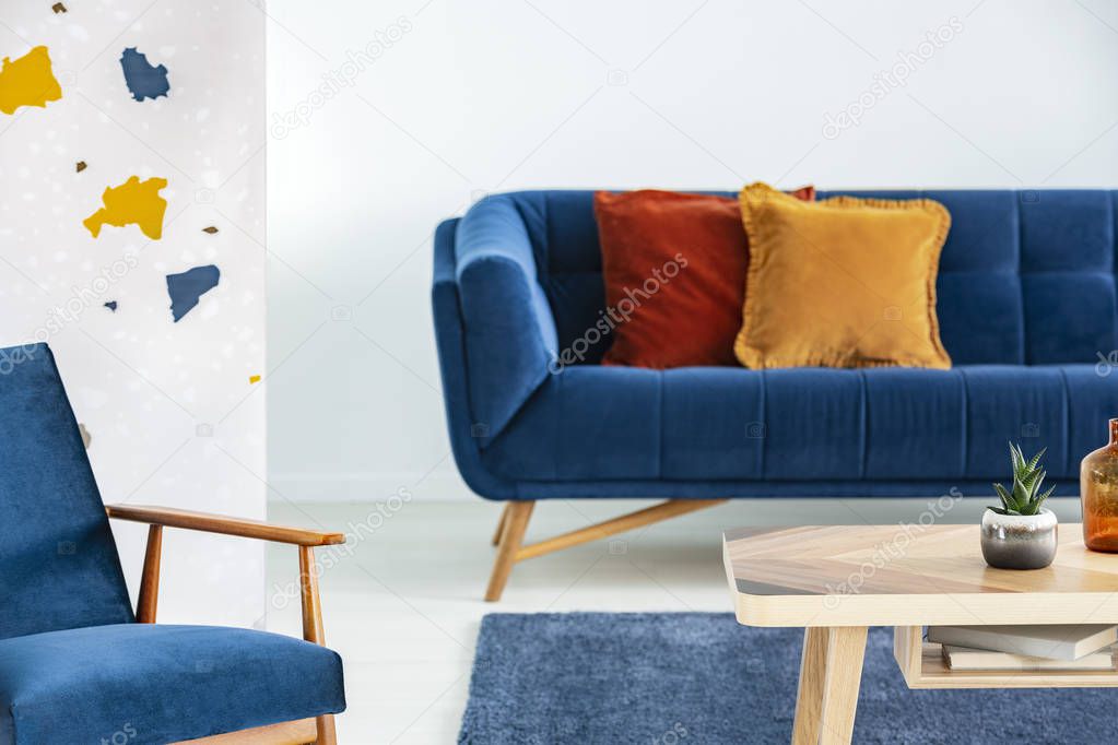 Armchair next to wooden table and blue sofa with pillows in modern living room interior. Real photo