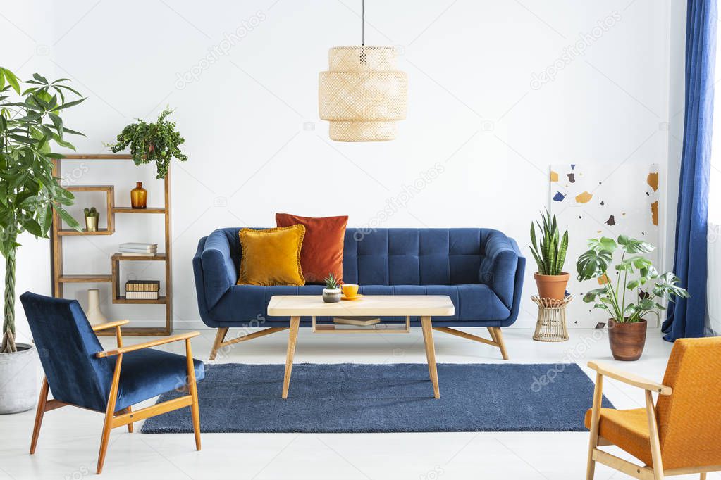 Retro armchairs with wooden frame and colorful pillows on a navy blue sofa in a vibrant living room interior with green plants. Real photo.