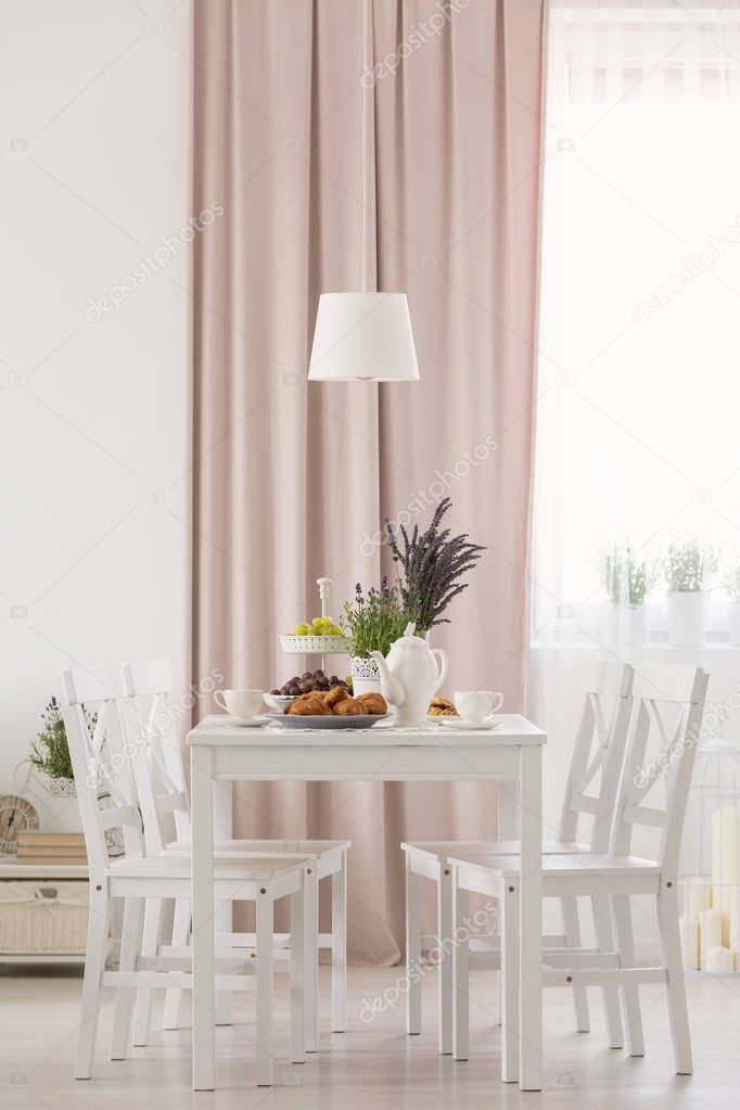White chairs at table with flowers under lamp in dining room interior with pink drapes. Real photo