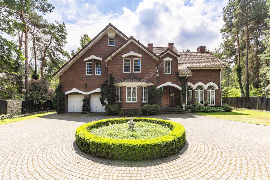 Front view of a driveway with a round garden and big, english style house in the background. Real photo