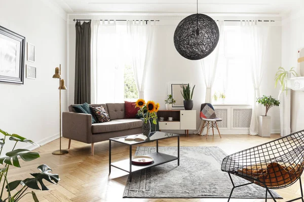 Sunflowers on table in bright flat interior with lamp next to settee near cabinet and chair. Real photo