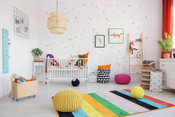 坐垫凳在斯堪婴儿卧室内的五颜六色的地毯上 配有台灯 摇篮和海报 真实照片 — 图库照片
