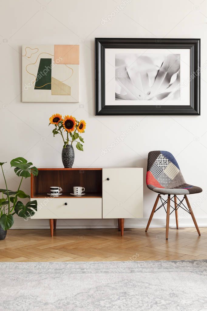 Framed photo and abstract art above sunflowers on an elegant cabinet and a patchwork chair in white living room interior.