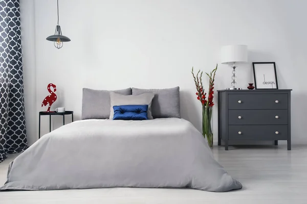 A monochromatic gray designer bedroom interior with a big bed and drawer cabinet and with contrasting accents of red gladiolas and blue quilted pillow. Real photo.