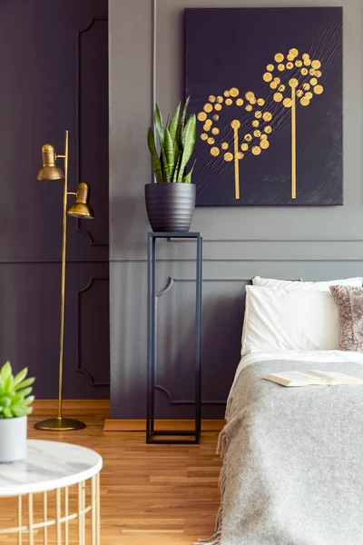 Plants, black poster and gold lamp in grey bedroom interior with book on bed. Real photo