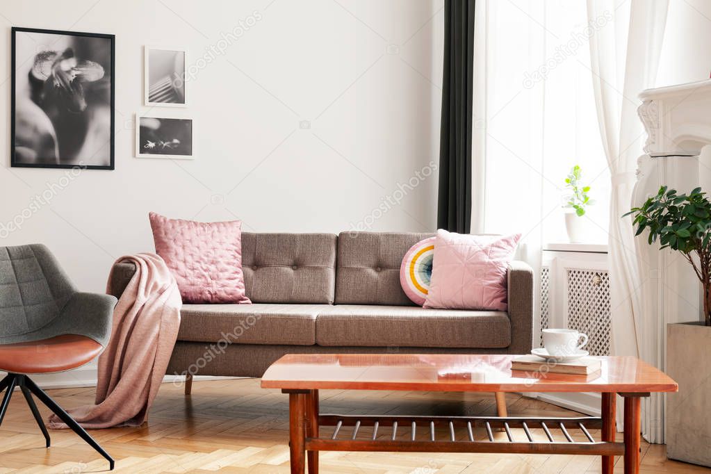 Pink pillows on sofa next to armchair in white living room interior with posters and table. Real photo