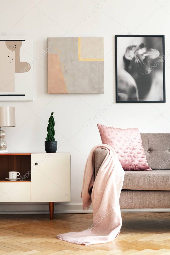 Real photo of bright sitting room interior with cupboard with fresh plant, herringbone parquet and couch with pastel pink blanket and pillow