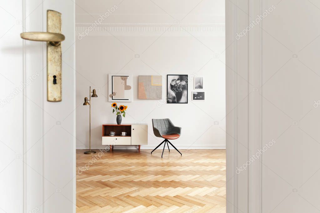 White living room interior with herringbone floor, gold lamp, gallery on wall and grey chair standing next to cupboard with sunflowers in real photo through the door