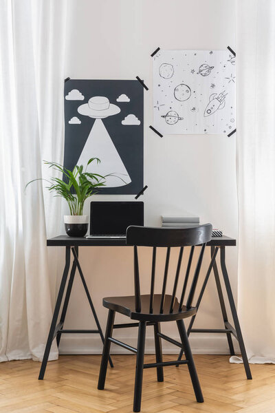 Black chair at desk with laptop and plant in white home office interior with posters. Real photo