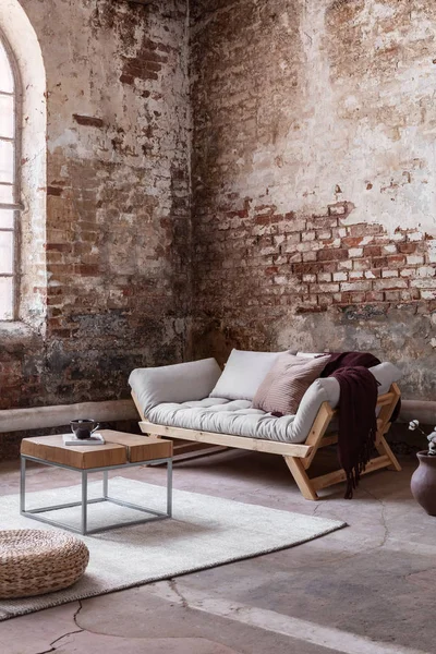 Pouf and wooden table on carpet in industrial loft interior with grey sofa against red brick wall. Real photo