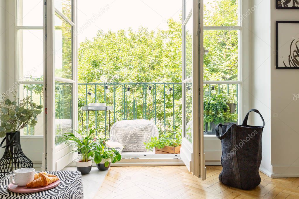 Open glass door from a living room interior into a city garden on a sunny balcony with green plants and comfy furniture