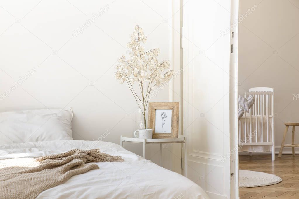 White metal bedside table with coffee mug, twig in glass vase and simple poster in frame placed by the bed in real photo of white bedroom interior