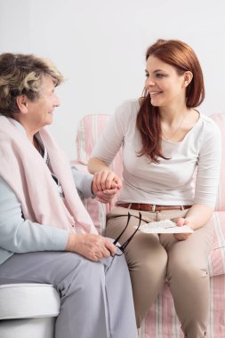 Happy caregiver talking with smiling senior woman while visit her at nursing house clipart