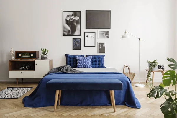 Upholstered bench on a wooden herringbone parquet in a bright and artistic bedroom interior with navy blue textiles