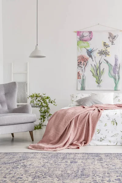 Cozy bedroom interior with a bed dressed in white and green sheets and peach blanket. Upholstered gray armchair beside the bed and painted fabric art on the back wall. Real photo.