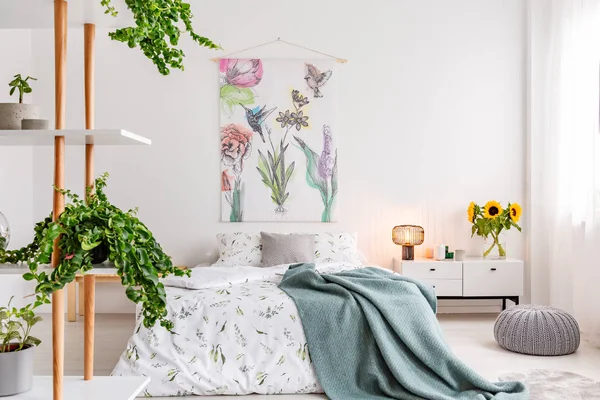 Green plants on shelves beside a bed dressed in white cotton bedding and teal blue blanket in a bright bedroom interior. Flowers and birds painted on the fabric above the bed. Real photo.