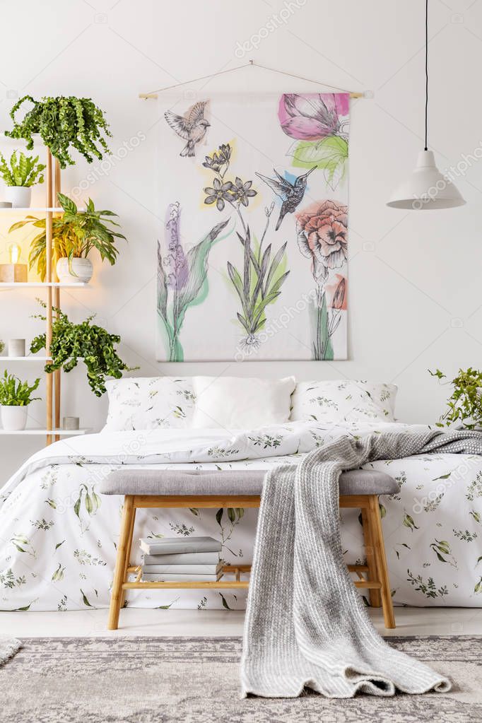 Urban jungle bedroom interior with a bed dressed in white and green bedding and painted art on the wall. Plants in pots beside the bed. Real photo.