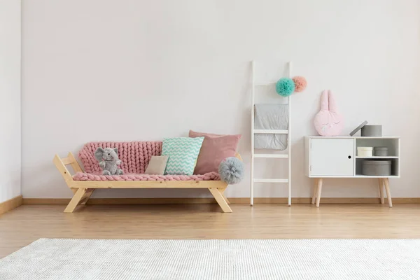 Comfortable sofa with pink blanket and colorful pillows next to white wooden ladder and shelf with round boxes, real photo with copy space on the white wall