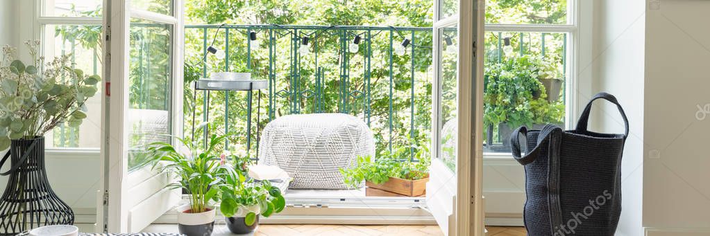 Panoramic view of open balcony door, black bag, green plants and light grey pouf