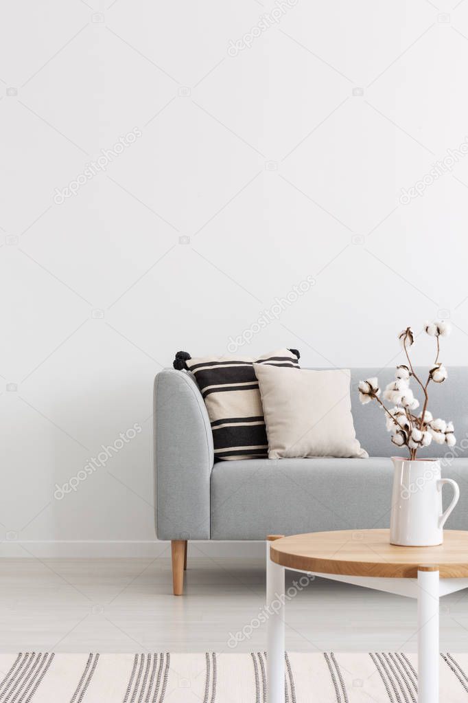 Flowers on wooden table in white minimal flat interior with pillows on grey sofa near rug. Real photo