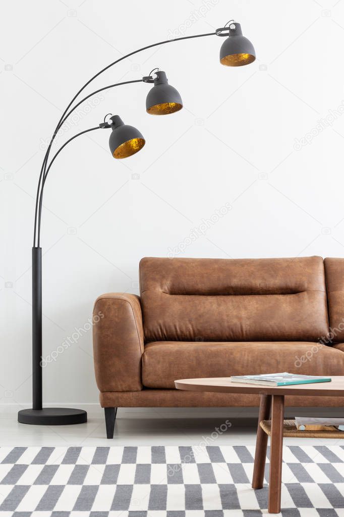 Vertical view of stylish black lamp and comfortable brown leather sofa in simple living room interior with coffee table and carpet