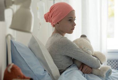 Oyuncak ayı tutan hastane yatağında oturup kanserli hasta çocuk