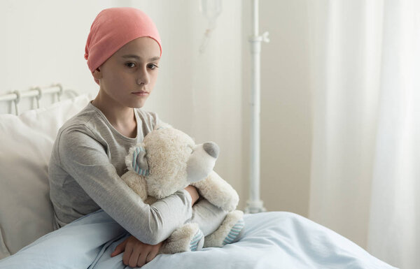 Sad sick girl with pink headscarf hugging plush toy in the hospital
