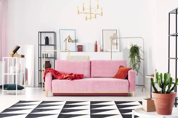 Metal shelves and abstract paintings behind powder pink couch in elegant white living room, real photo