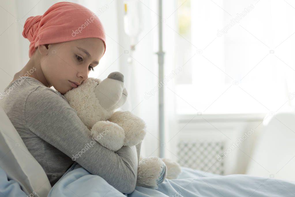 Weak girl with cancer wearing pink headscarf and hugging teddy bear 