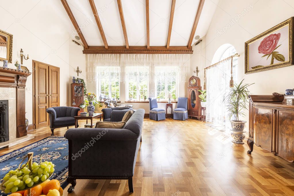 A view of a luxurious high ceiling living room interior with wooden floor, sunny windows and classic settee, armchairs and cabinet. Empty space on the floor. Real photo.