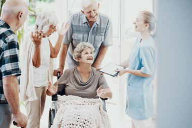 Gülümseyen arkadaşları ve felçli kadın kıdemli tekerlekli sandalyede destekleyen hemşire