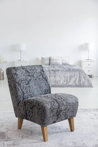 Grey armchair on carpet in white bedroom interior with pillows on bed between lamps. Real photo