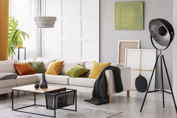 White elegant corner sofa with orange green and yellow pillows in stylish living room interior with modern coffee table and industrial lamp