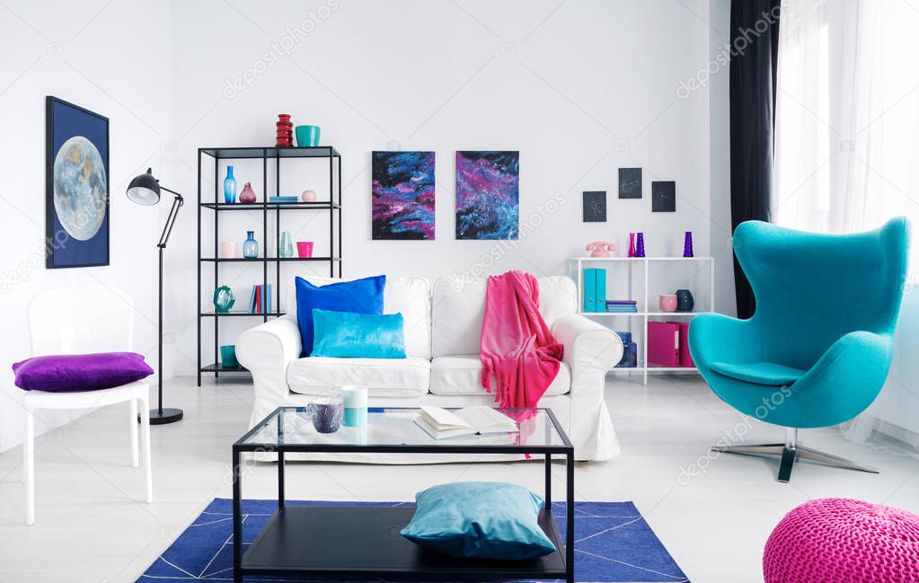 Stylish white living room with colorful accessories, white couch and metal coffee table in the middle next to blue egg chair, real photo