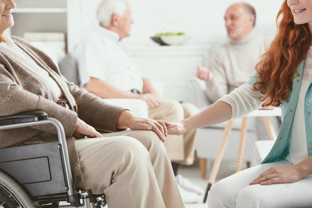Friendly young nurse supporting paralyzed senior woman on wheelchair in nursing house
