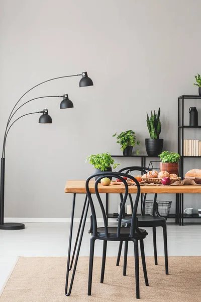 Lamp and plants in grey dining room interior with black chairs at wooden table with food. Real photo