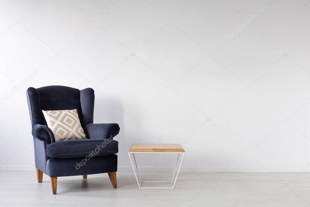 Patterned white and gold pillow on elegant dark blue armchair in stylish white interior with coffee table, real photo