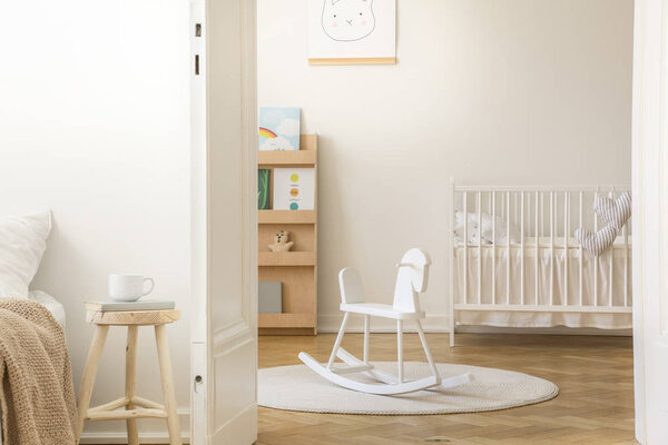 Scandinavian nursery with white wooden crib, round carpet and rocking horse in the middle, real photo with copy space