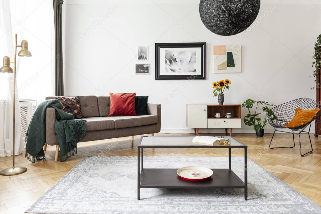 Table on carpet in white apartment interior with sofa, armchair and posters above cabinet. Real photo