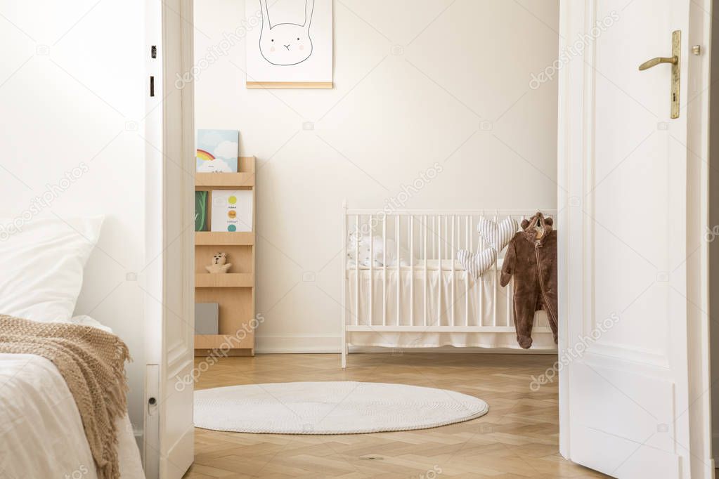 Stylish bedroom interior with open door to scandinavian nursery with white crib, poster on the wall and carpet on the floor, real photo